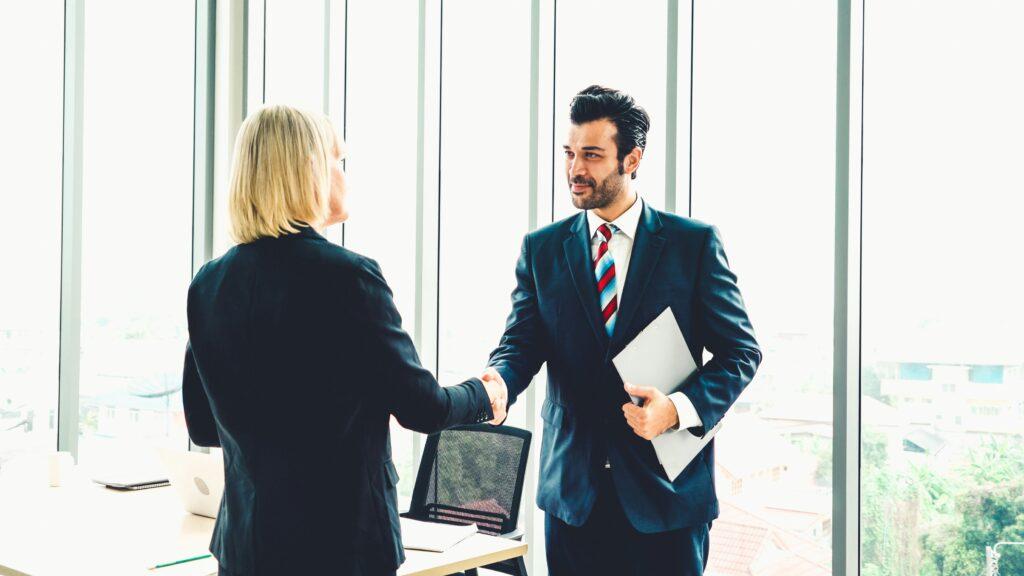 A business professional in a suit holding documents shakes hands with another person in an office with large windows overlooking a bright cityscape, suggesting a successful meeting or agreement.