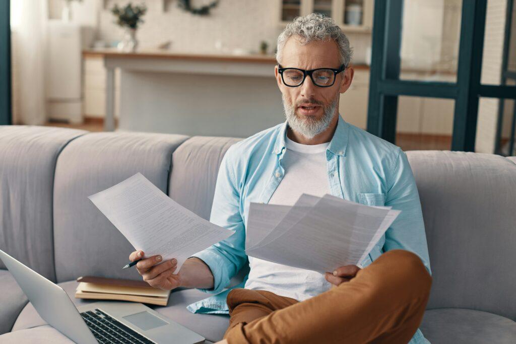 A middle-aged man with gray hair and glasses sitting on a sofa, holding multiple documents in both hands while reviewing them, with an open laptop, folders, and a pen beside him in a home setting.