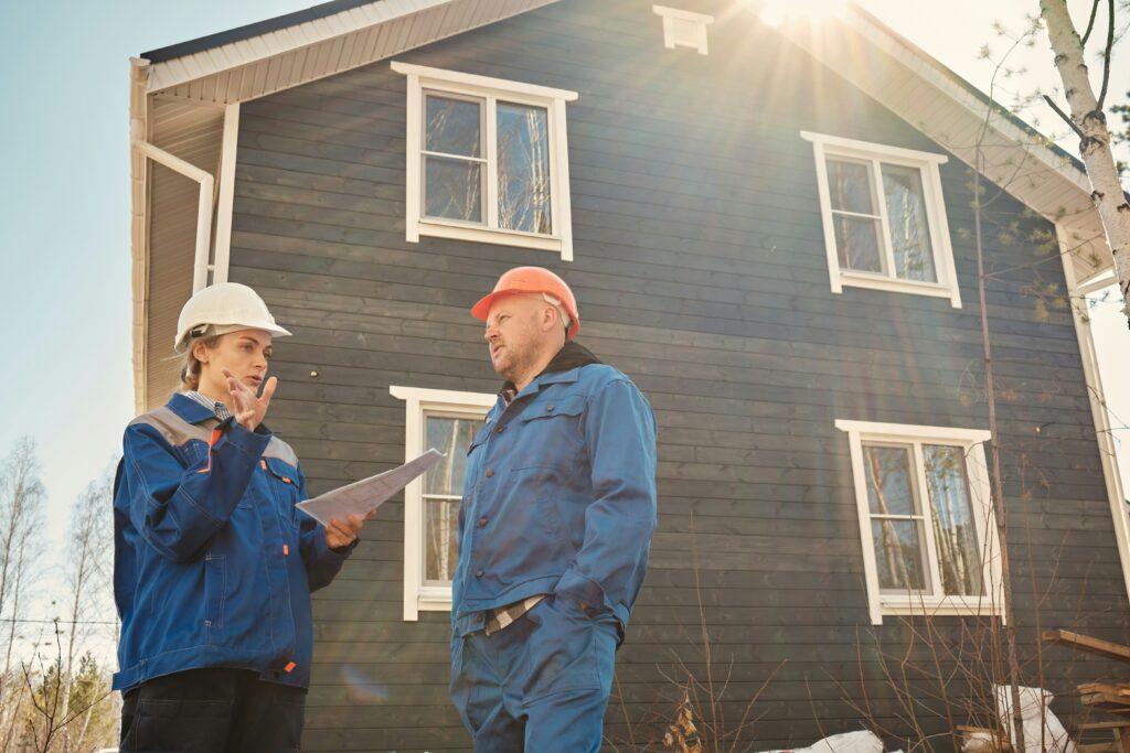 Construction worker and site inspector wearing hard hats discuss plans while reviewing documents outside a newly built two-story house in daylight.