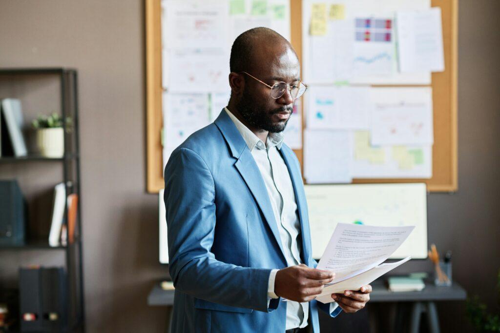 Business professional in a blue suit reviews printed documents while standing in an office with charts and notes pinned on a bulletin board in the background.