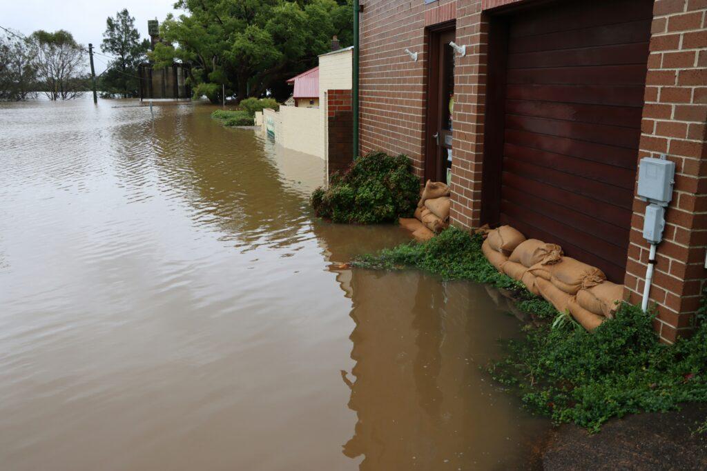Flooded residential street with muddy brown water reaching the entrances of brick houses, sandbags stacked in front of a garage and doorway as a temporary barrier, vegetation partially submerged along the building, and trees and utility poles visible in the distance under overcast skies.