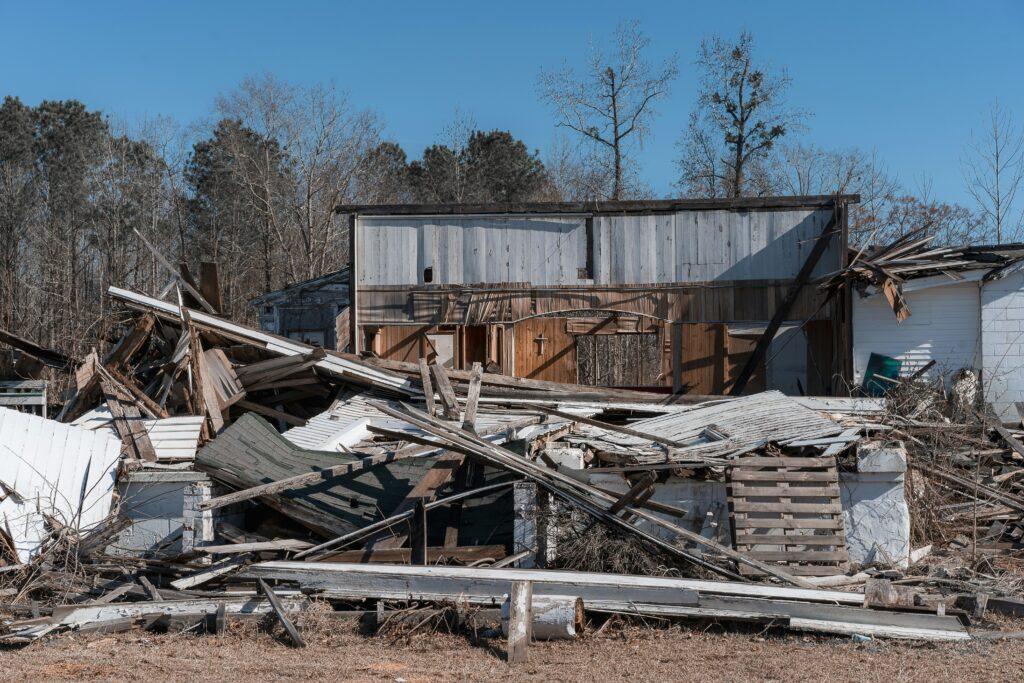 Collapsed wooden building with extensive structural damage, broken walls, fallen roof panels, and scattered debris across the ground in a rural outdoor setting.