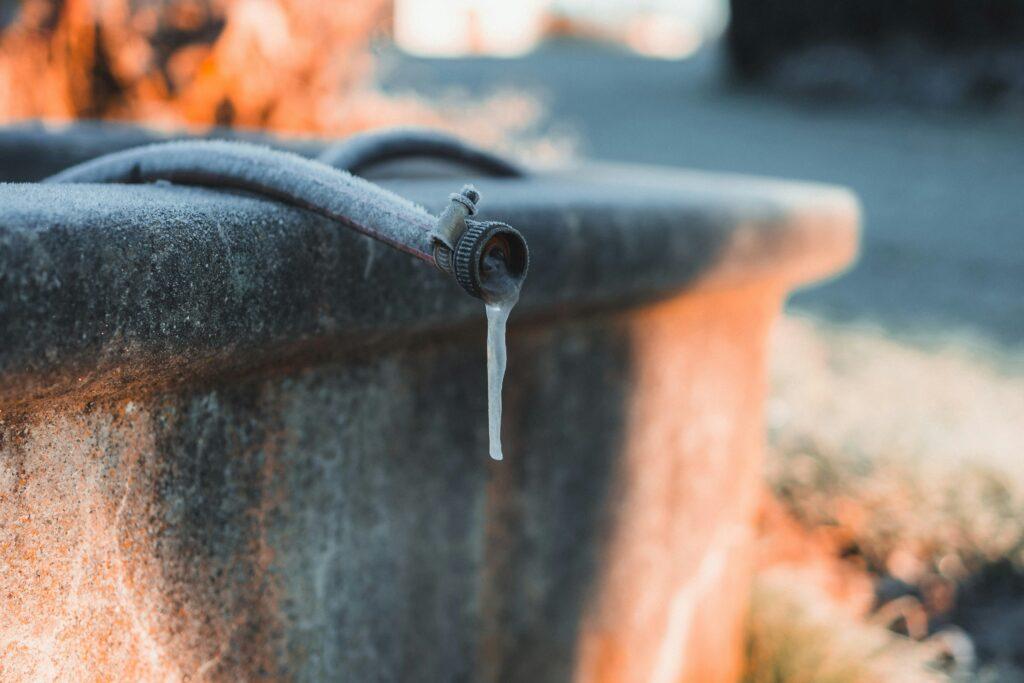 Frozen outdoor faucet with a small icicle hanging from the spout, attached to a metal surface in cold weather conditions